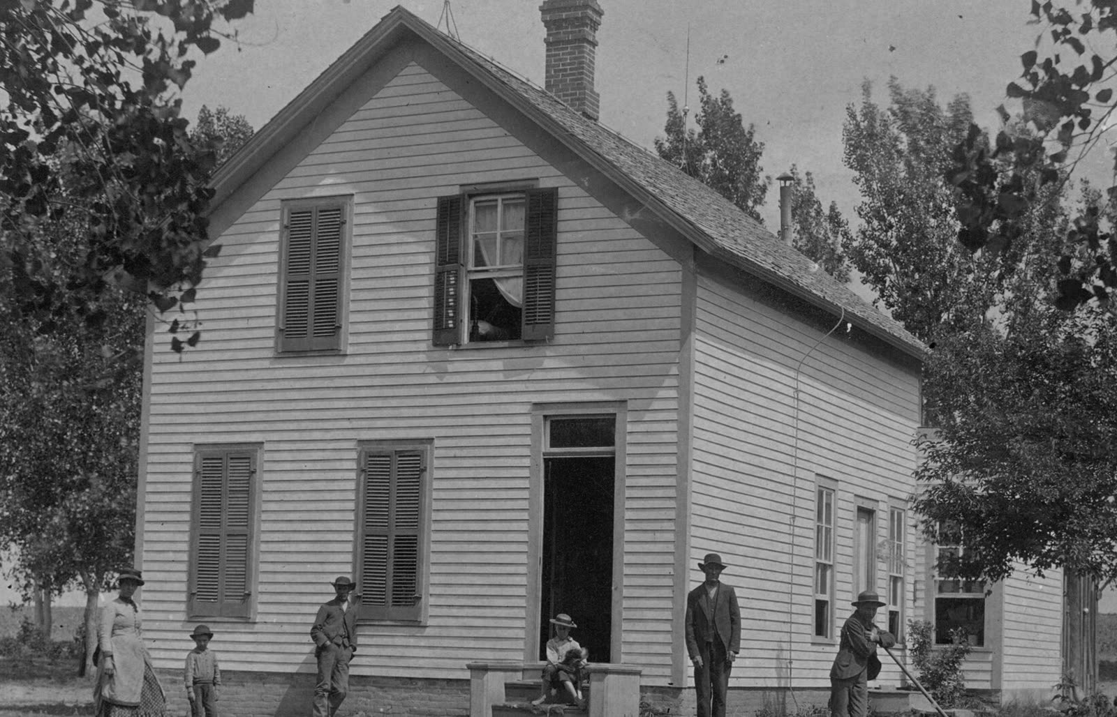 Old photograph: a plain white gabled house with shutters, a family standing in the yard in front of it, about 1885