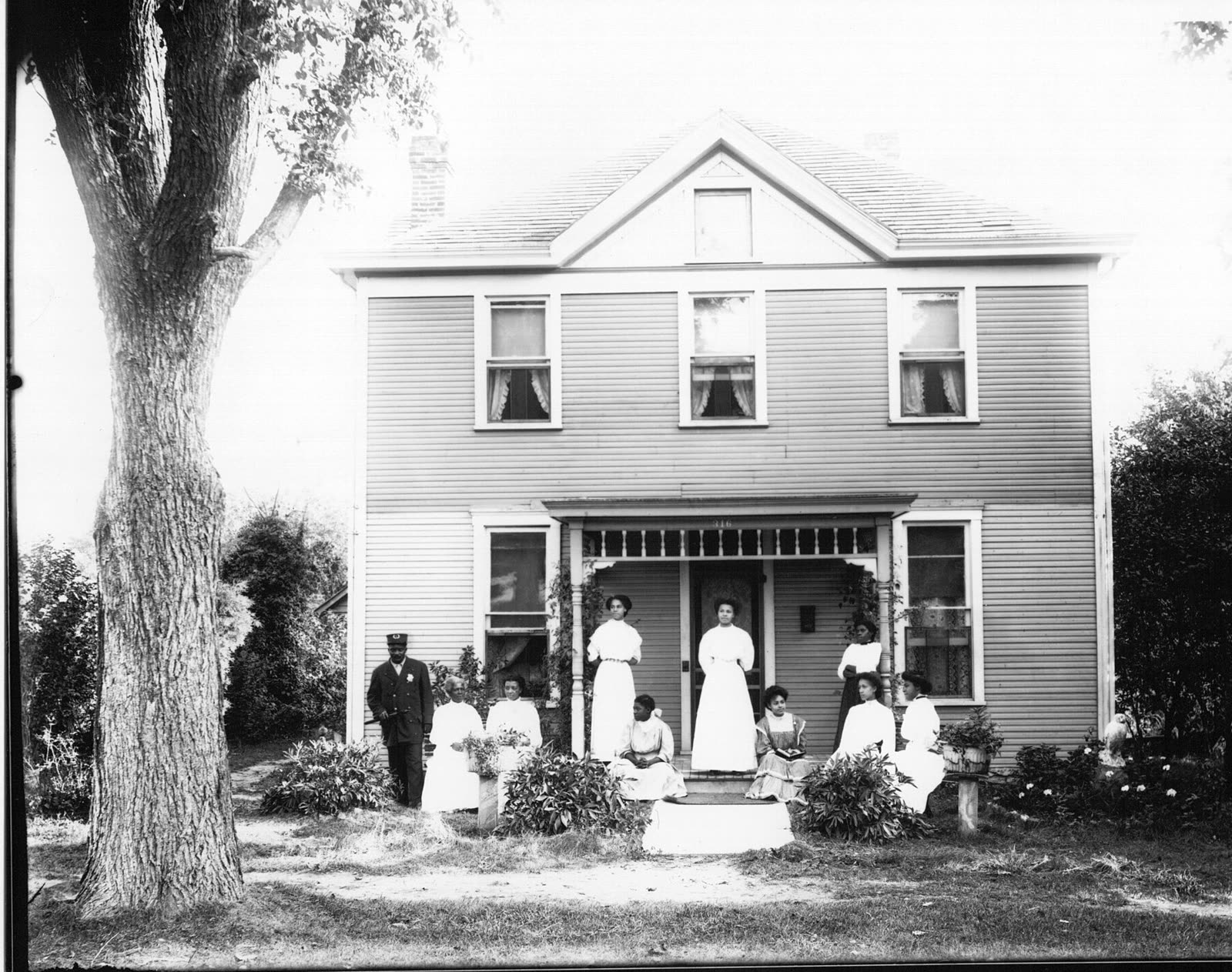 Old photograph: a family of eight standing on the lawn and porch steps of a two-storey clapboard house, 1908