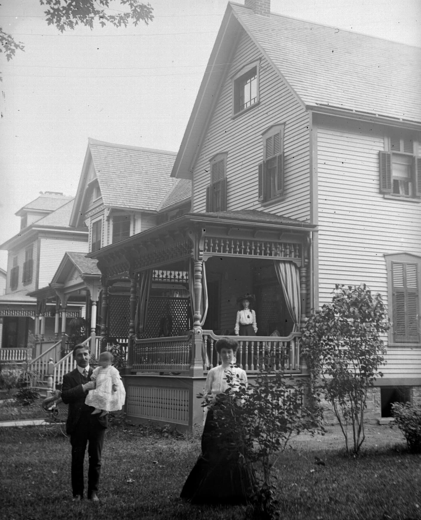 Old black-and-white photograph: a tall clapboard house with a porch, a man holding a baby and a woman standing on the lawn in front of it