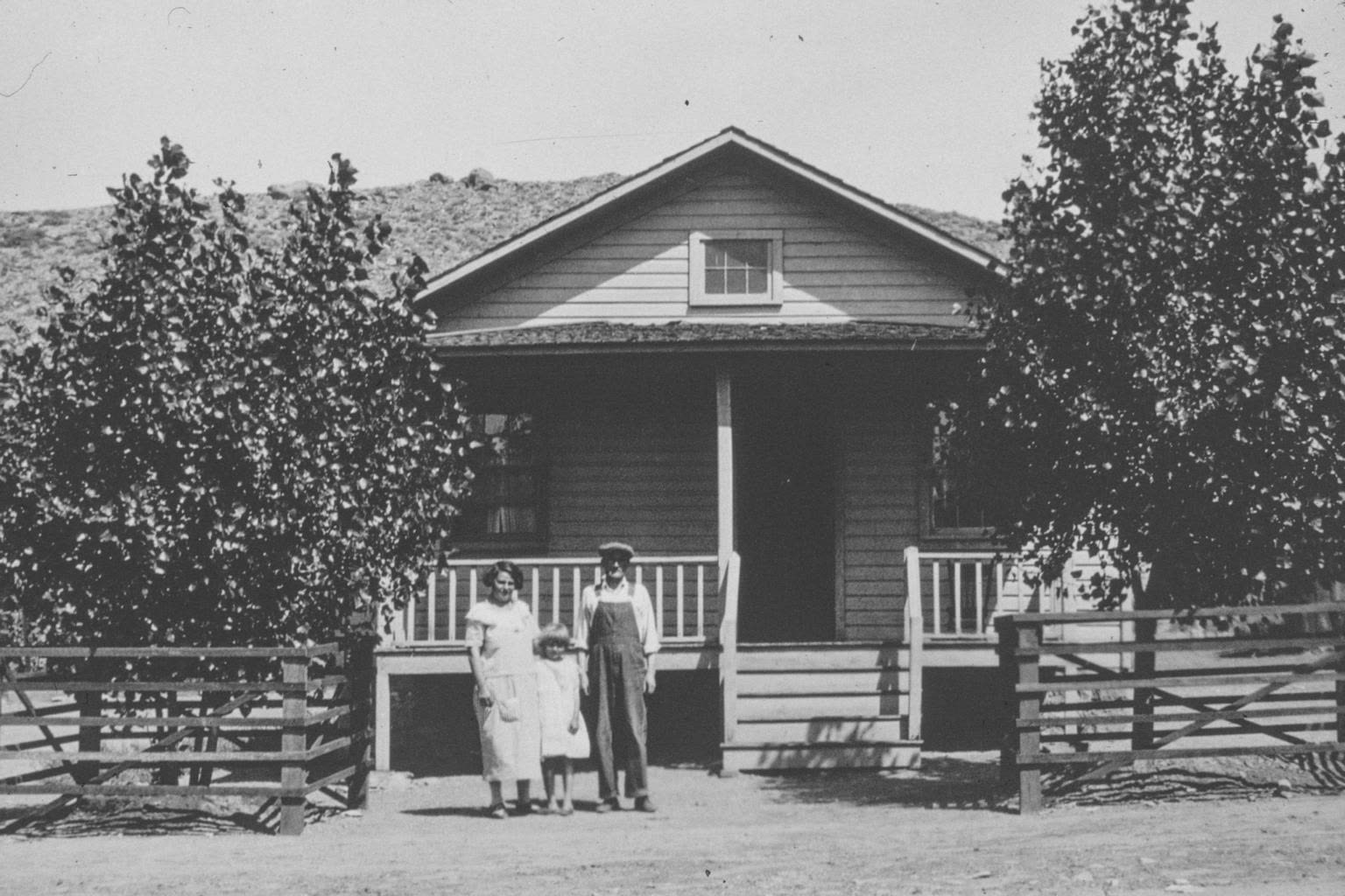 Old photograph: a man, a woman and two children in front of the porch of a small house behind a wooden fence, 1924